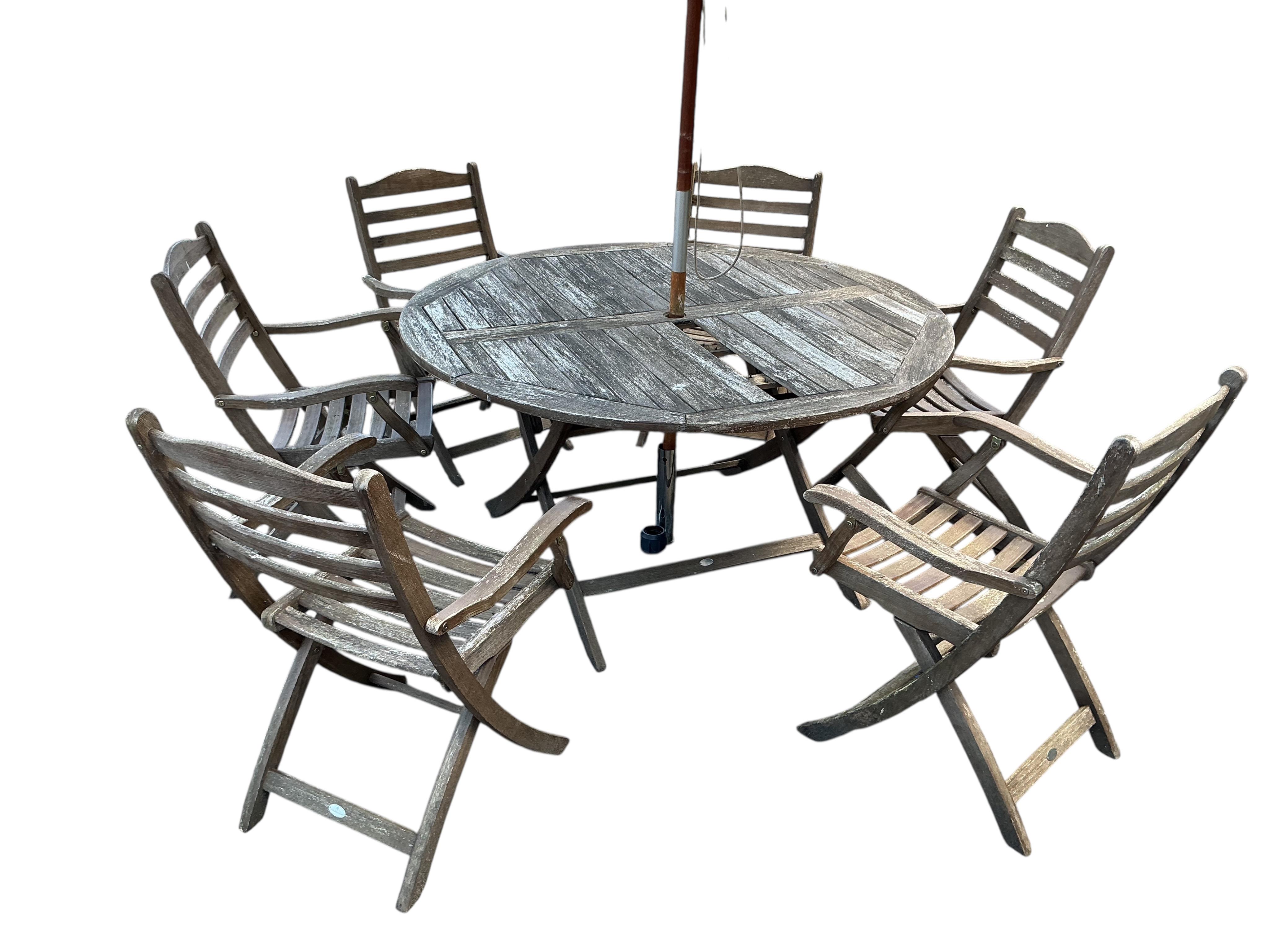 A circular weatherd teak garden table, diameter 140cm, height 74cm with six chairs by Alexander Rose, and a parasol on carved stone base (table in need of repair)